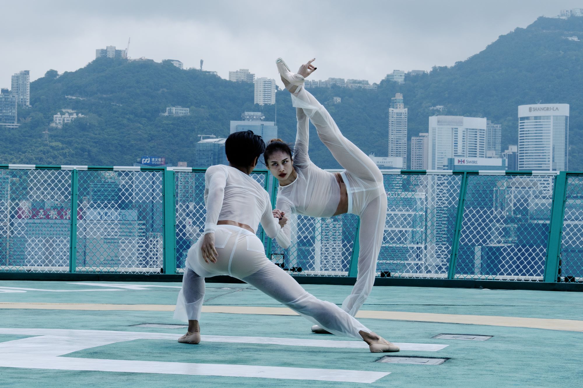 Two women in white outfits performing acrobatics with Avenue of Stars, Hong Kong in the background